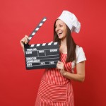 Housewife female chef cook or baker in striped apron, white t-shirt, toque chefs hat isolated on red wall background. Woman holding classic black film making clapperboard. Mock up copy space concept